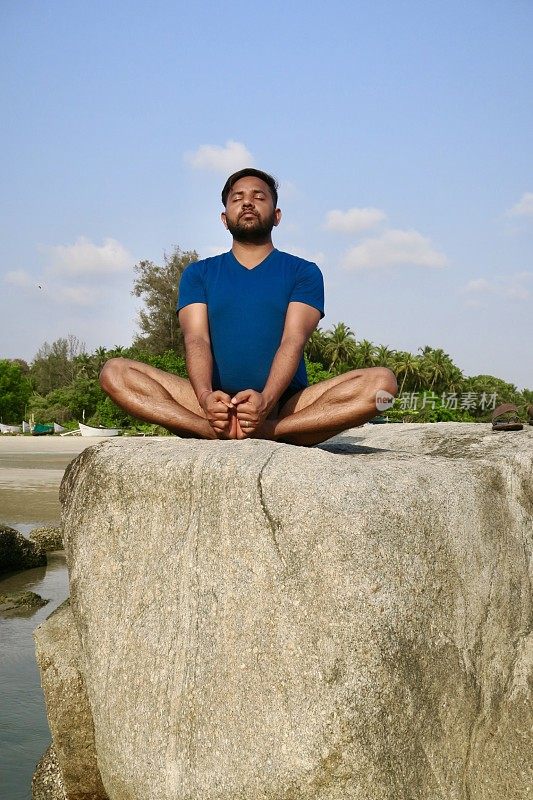 印度男子在岩石上练习蝴蝶/鞋匠姿势(Baddha Konasana)瑜伽姿势，水边，Palolem海滩，果阿，印度